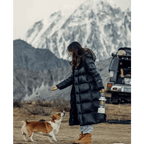Femme en manteau noir interagissant avec un chien devant des montagnes enneigées, près d'un véhicule tout-terrain. Aventure, nature, randonnée.