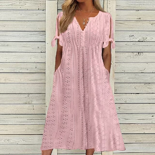 Robe longue rose en dentelle ajourée, manches courtes nouées, col en V, devant un mur en bois clair. Mode féminine élégante et estivale.