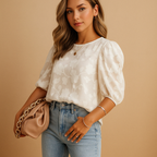 Jeune femme portant un chemisier blanc à motifs floraux, tenant un sac à main beige avec chaîne dorée, posant devant un fond beige.