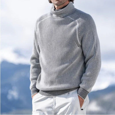 Homme portant un pull gris en laine, col roulé, dans un paysage montagneux enneigé. Mode hiver, vêtements chauds, style décontracté.
