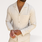 Homme portant une chemise en lin beige, manches longues, col ouvert, avec un collier doré. Mode masculine élégante et décontractée.