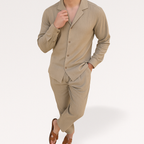 Homme en costume beige élégant, chemise à col ouvert, pantalon assorti, sandales marron, pose décontractée. Mode masculine moderne, style chic.