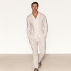 Homme portant un ensemble en lin beige, chemise à manches longues et pantalon assorti, marchant sur un fond neutre. Mode masculine élégante et décontractée.