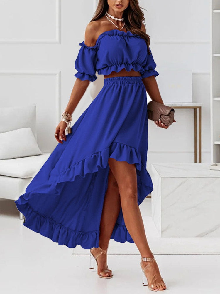 Robe bleue élégante à volants, épaules dénudées, jupe longue asymétrique, portée avec des sandales transparentes et un sac à main beige.