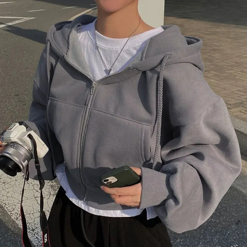 Personne portant un sweat à capuche gris, tenant un appareil photo argenté et un smartphone vert. Mode décontractée, rue, photographie urbaine.