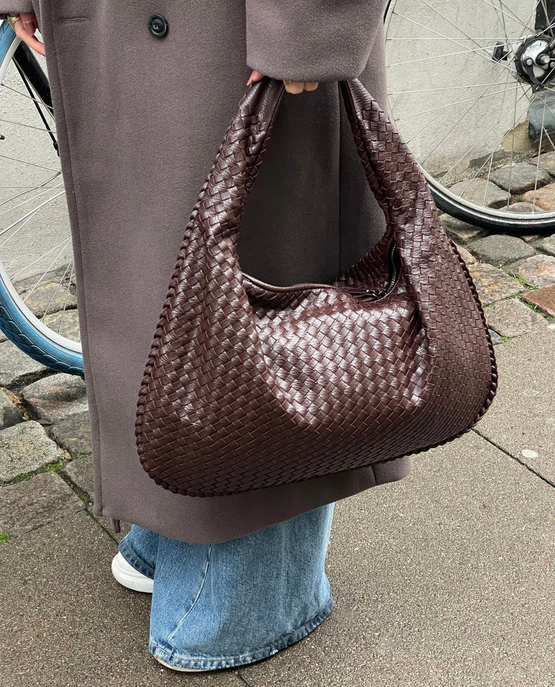Sac à main en cuir tressé marron porté par une personne en manteau gris et jean, avec une roue de vélo en arrière-plan. Mode urbaine élégante.
