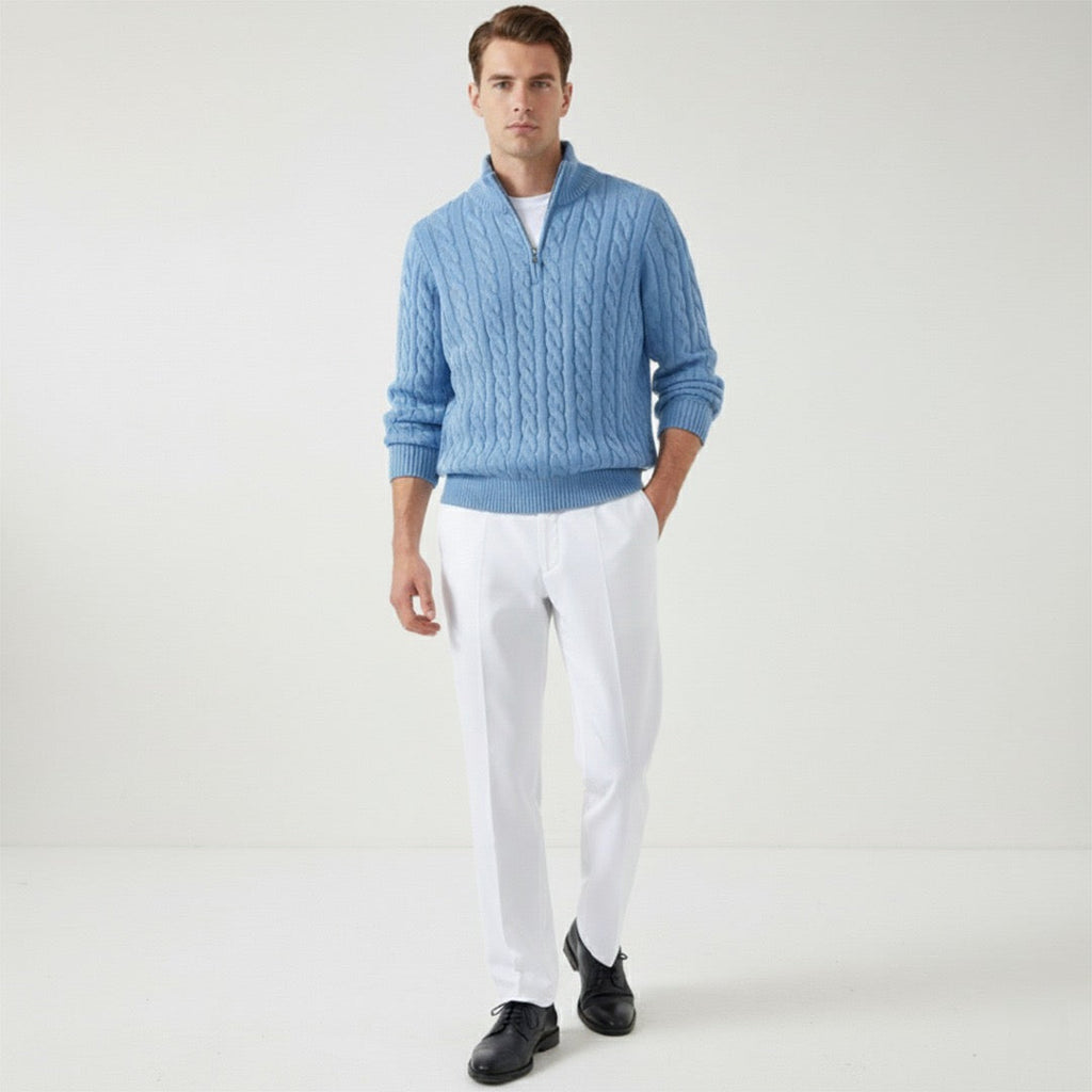 Homme portant un pull bleu en laine torsadée, pantalon blanc et chaussures noires, posant dans un studio minimaliste. Mode masculine élégante.