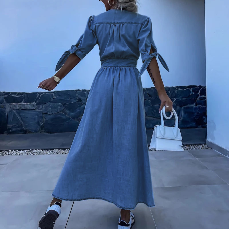 Femme en robe longue en jean bleu, manches nouées, tenant un sac blanc, debout sur une terrasse moderne. Mode estivale élégante et décontractée.