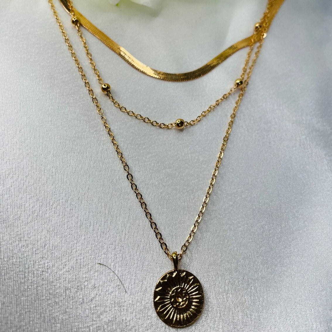 Collier en or à trois rangs avec pendentif soleil, chaîne perlée et maille plate, sur fond blanc. Bijoux élégants, tendance mode, accessoires féminins.