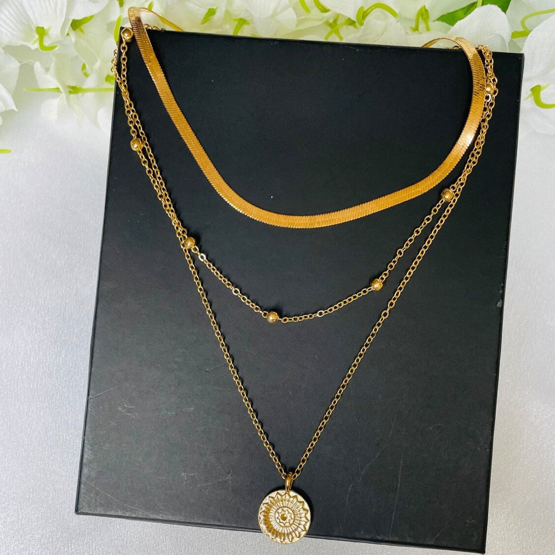 Collier multi-rangs doré avec pendentif rond floral, chaînes fines et perles, sur fond noir. Bijoux élégants, tendance mode féminine.