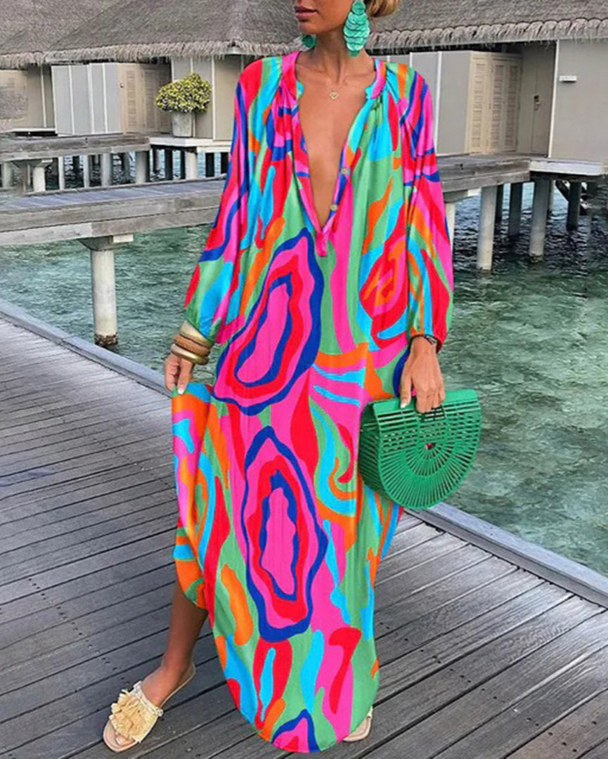 Robe longue colorée à motifs abstraits, femme tenant un sac vert en osier, debout sur une terrasse en bois près de l'eau, style estival chic.