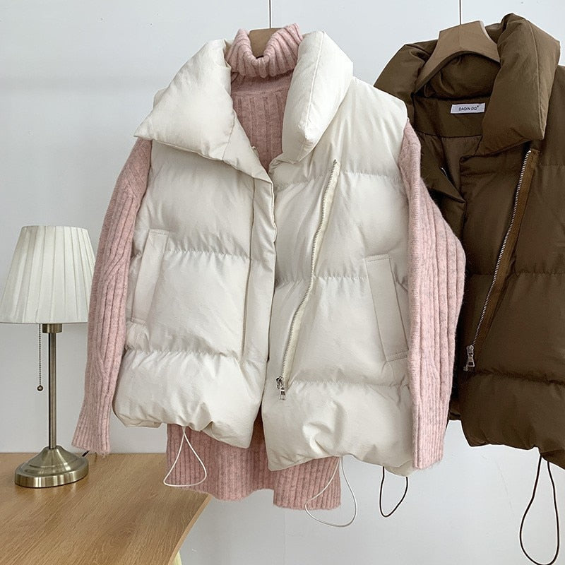 Gilet matelassé blanc suspendu sur un pull rose en tricot, à côté d'un gilet marron, sur un fond de lampe et table en bois. Mode hiver tendance.