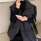 Femme en manteau noir matelassé, tenant un smartphone blanc, assise sur un canapé beige. Sac à chaîne dorée visible. Mode hiver élégante.