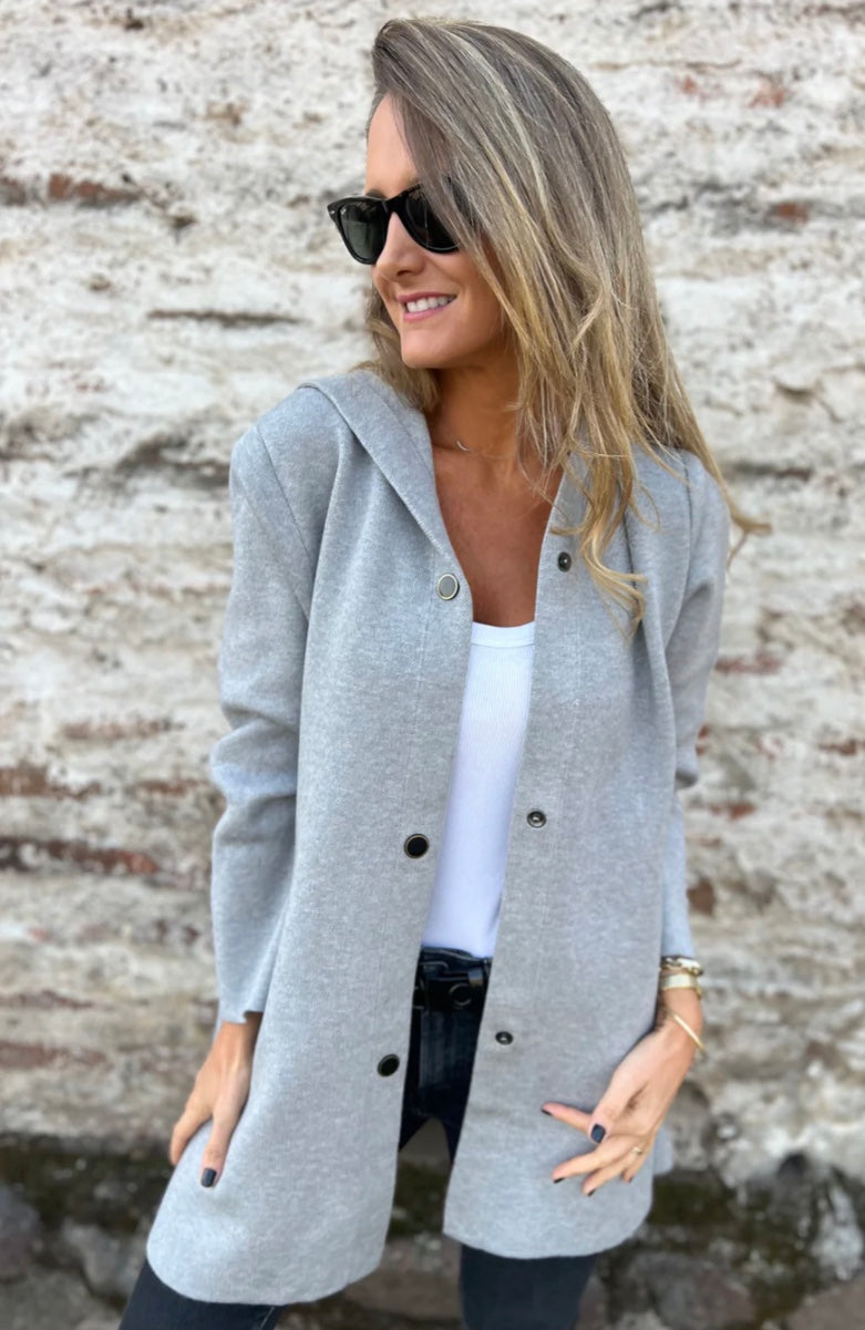 Femme souriante portant un manteau gris élégant, lunettes de soleil noires, devant un mur en pierre. Mode automne, style décontracté chic.
