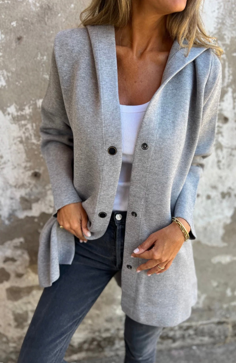 Femme portant un manteau gris élégant, chemise blanche et jeans, posant contre un mur texturé. Mode automne, style décontracté chic.