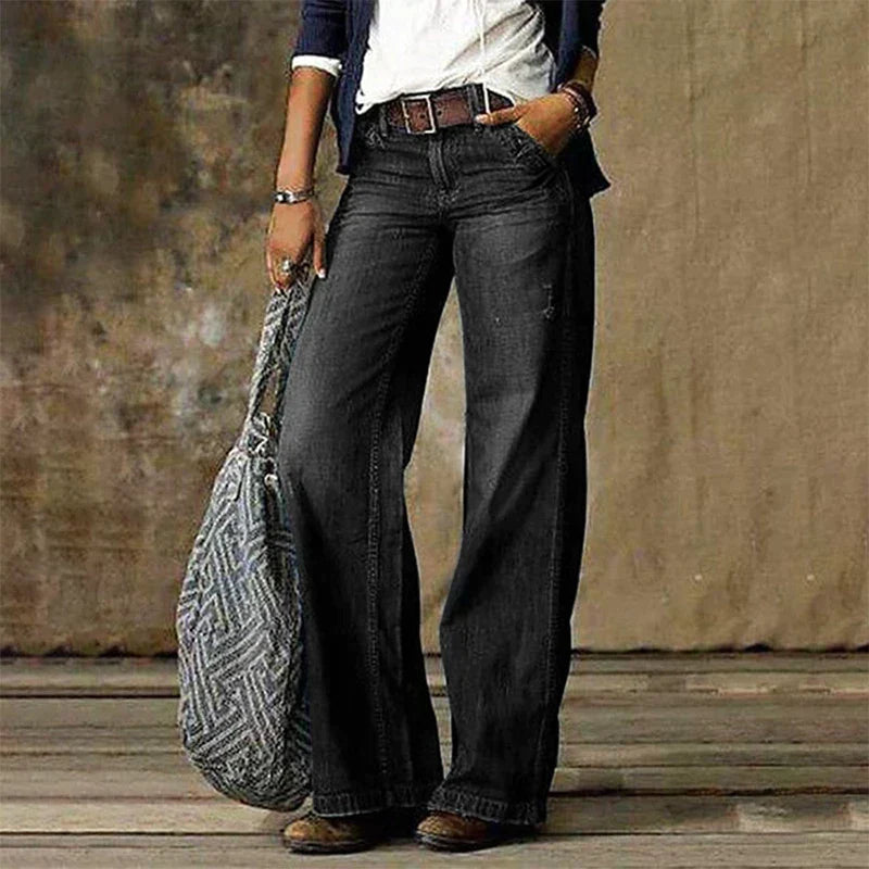 Femme portant un jean large noir, chemise blanche et veste bleue, tenant un sac à motif géométrique. Mode décontractée, style vintage.
