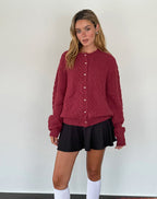 Jeune femme portant un cardigan rouge texturé avec boutons, jupe noire et chaussettes blanches. Mode automne-hiver, style décontracté et élégant.