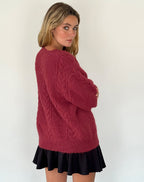Jeune femme portant un pull en laine rouge bordeaux texturé, jupe noire plissée, posant contre un mur blanc. Mode automne-hiver, style décontracté.