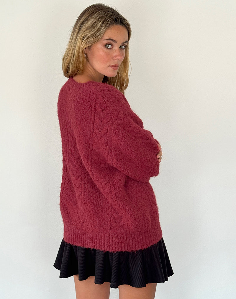 Jeune femme portant un pull en laine rouge bordeaux texturé, jupe noire plissée, posant contre un mur blanc. Mode automne-hiver, style décontracté.