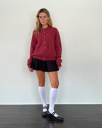 Jeune femme portant un cardigan rouge texturé, jupe noire, chaussettes hautes blanches et chaussures noires, posant devant un mur blanc.