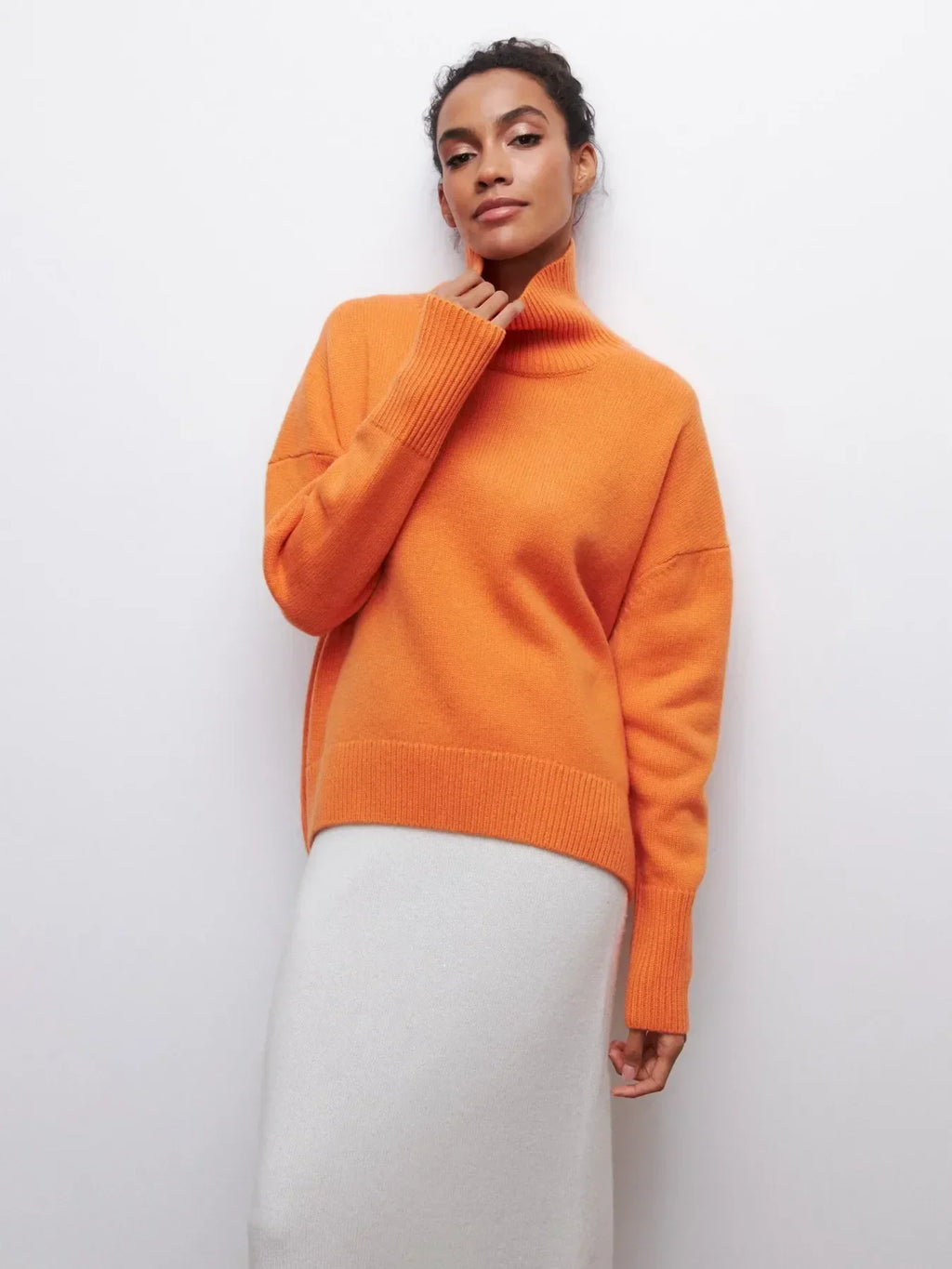 Femme portant un pull orange à col roulé et une jupe blanche, posant contre un mur blanc. Mode automne-hiver, style élégant et moderne.