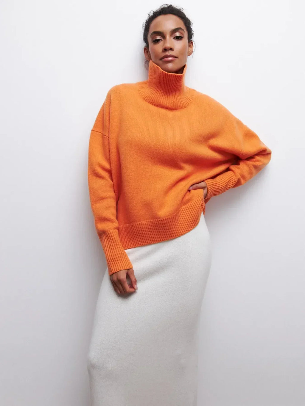Femme portant un pull orange à col roulé et une jupe blanche, posant contre un mur blanc. Mode automne-hiver, style élégant et moderne.