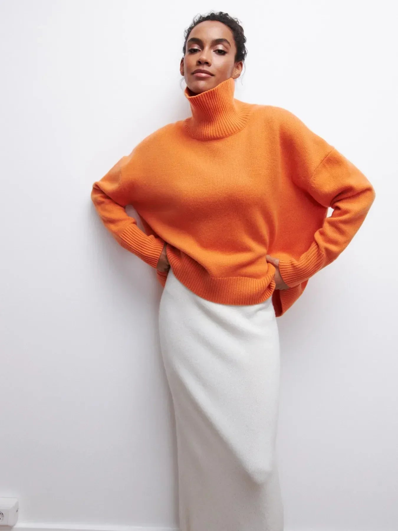 Femme portant un pull orange à col roulé et une jupe longue blanche, posant contre un mur blanc. Mode automne-hiver, style élégant et moderne.
