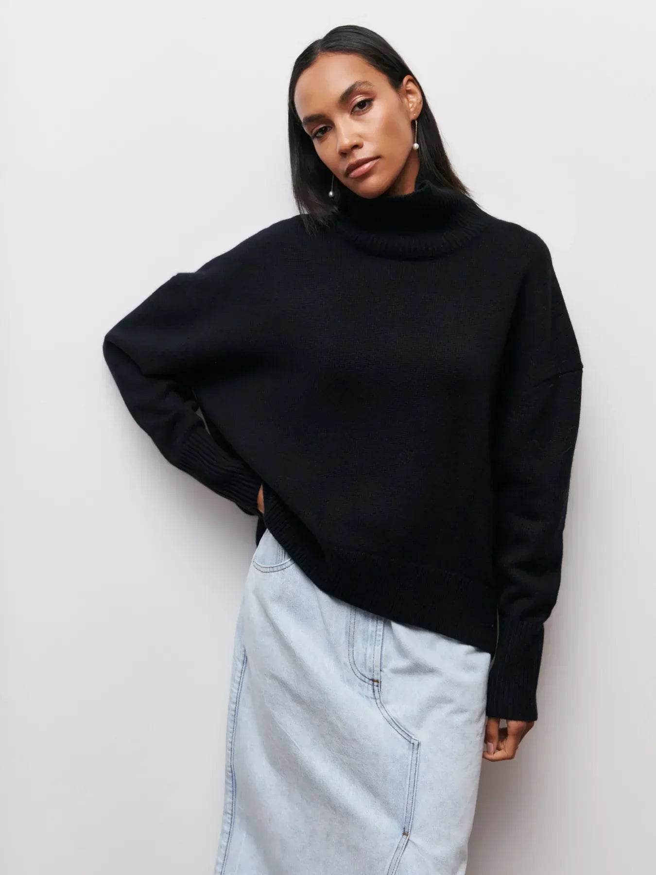 Femme portant un pull noir à col roulé et une jupe en jean clair, posant contre un mur blanc. Mode automne-hiver, style décontracté chic.