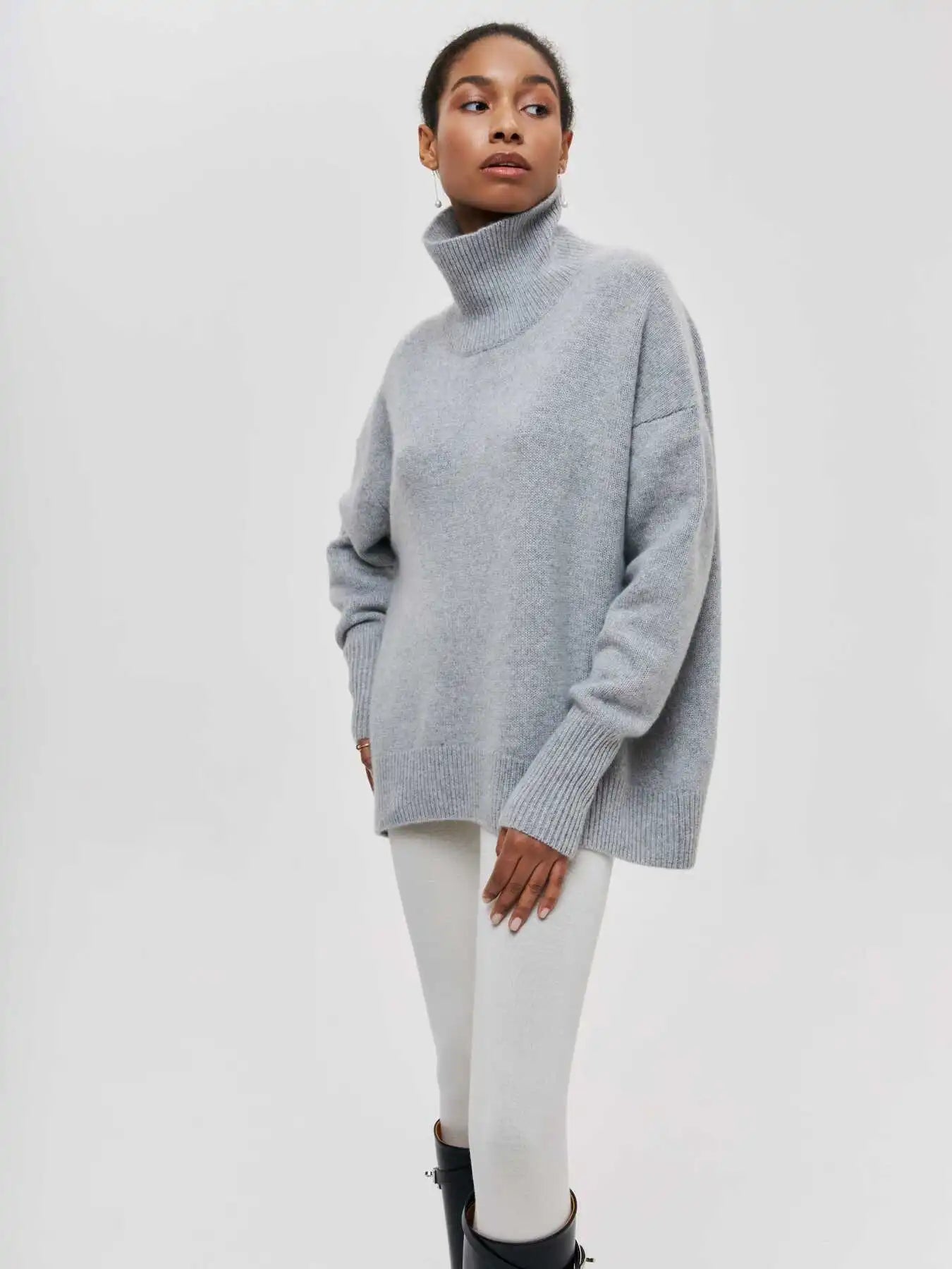 Femme portant un pull gris à col roulé, manches longues, associé à un legging blanc et des bottes noires. Mode automne-hiver élégante et confortable.