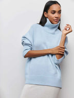 Femme portant un pull bleu clair à col roulé, manches longues, posant contre un mur blanc. Mode automne-hiver, style décontracté et élégant.
