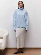 Femme portant un pull bleu à col roulé et une jupe longue beige, posant dans un intérieur minimaliste. Mode automne-hiver élégante et moderne.
