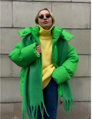 Femme en manteau vert vif, pull jaune, écharpe verte, lunettes de soleil rondes, posant devant un mur en pierre. Mode hiver tendance.