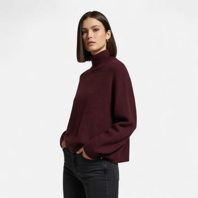 Femme portant un pull bordeaux à col roulé, manches longues, posant avec les mains dans les poches d'un jean noir. Mode automne-hiver élégante.