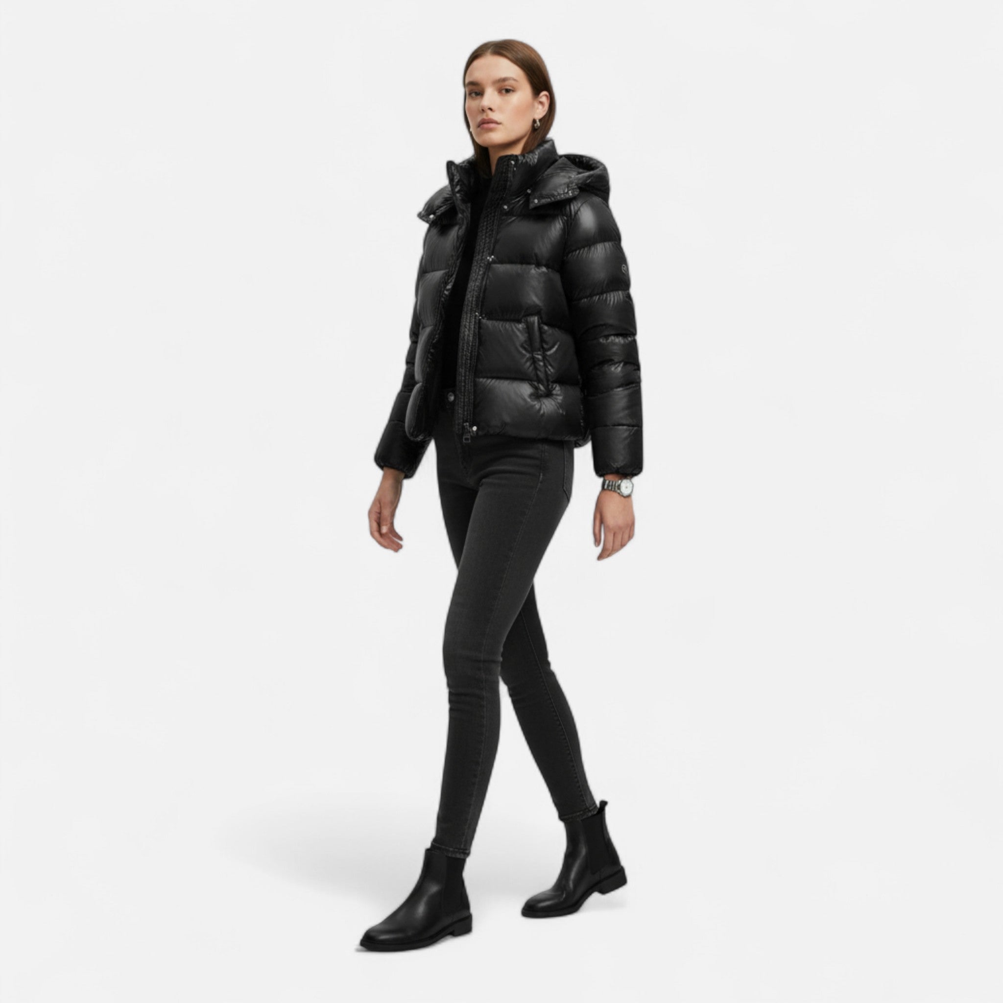 Femme portant une doudoune noire élégante, pantalon slim noir et bottines en cuir, marchant sur fond blanc. Mode hiver chic, tendance urbaine.