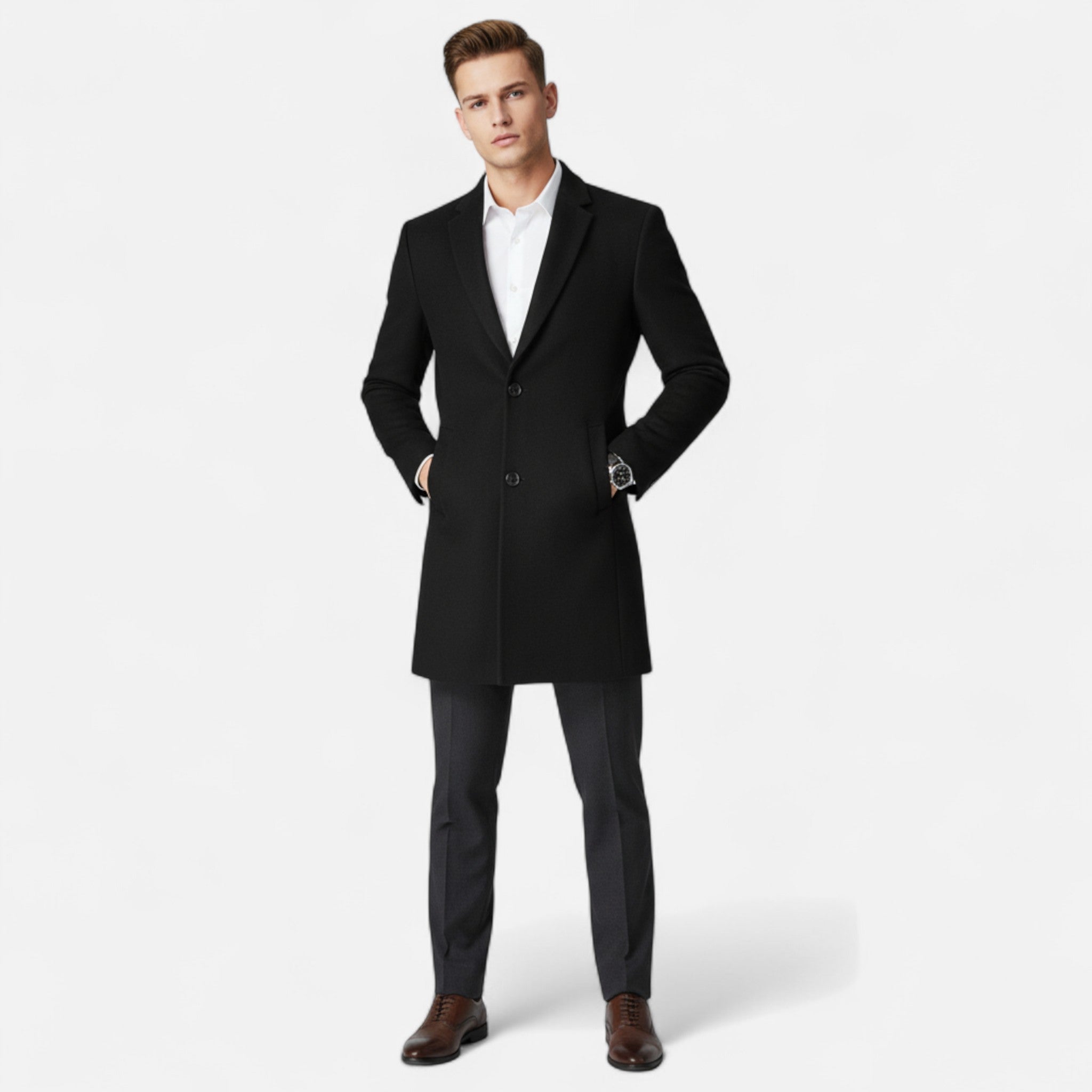 Homme en manteau noir élégant, chemise blanche, pantalon gris et chaussures marron, posant sur fond blanc. Mode masculine chic et moderne.