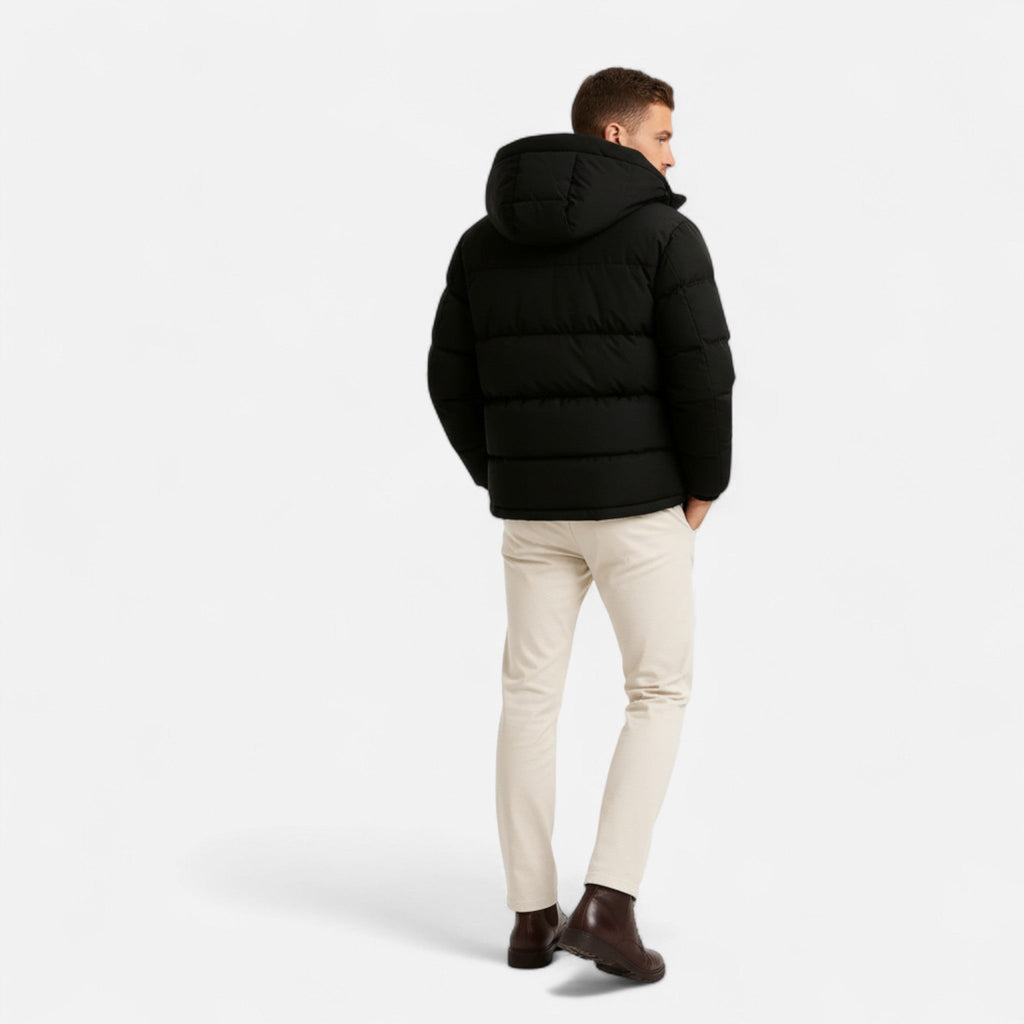 Homme portant une doudoune noire avec capuche, pantalon beige et bottines marron, vu de dos sur fond blanc. Mode hiver, tenue décontractée.