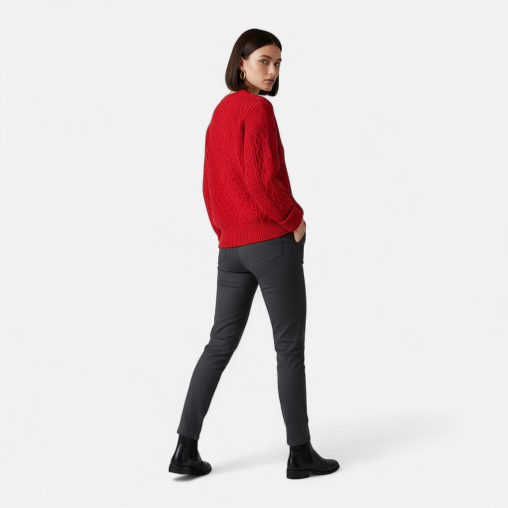 Femme portant un pull rouge en laine, pantalon gris et bottines noires, posant sur fond blanc. Mode automne-hiver, style décontracté et élégant.