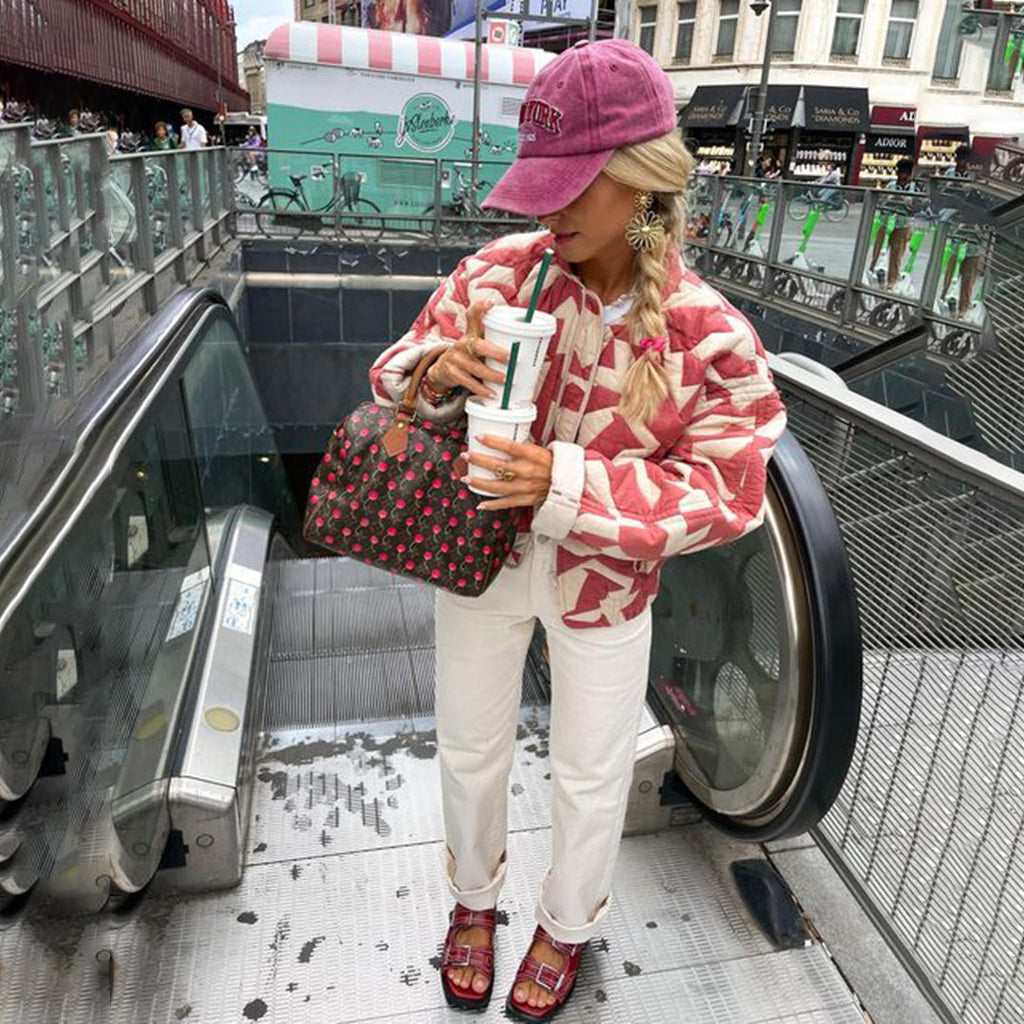 Femme à la mode portant une veste rose et blanche, casquette violette, tenant un sac à motifs et un café, sur un escalator urbain.