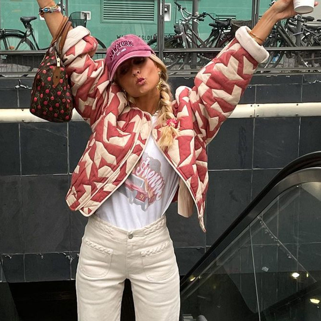 Femme en veste matelassée rouge et blanche, casquette rose, tenant un sac à main et un café, posant devant des vélos garés. Mode urbaine tendance.