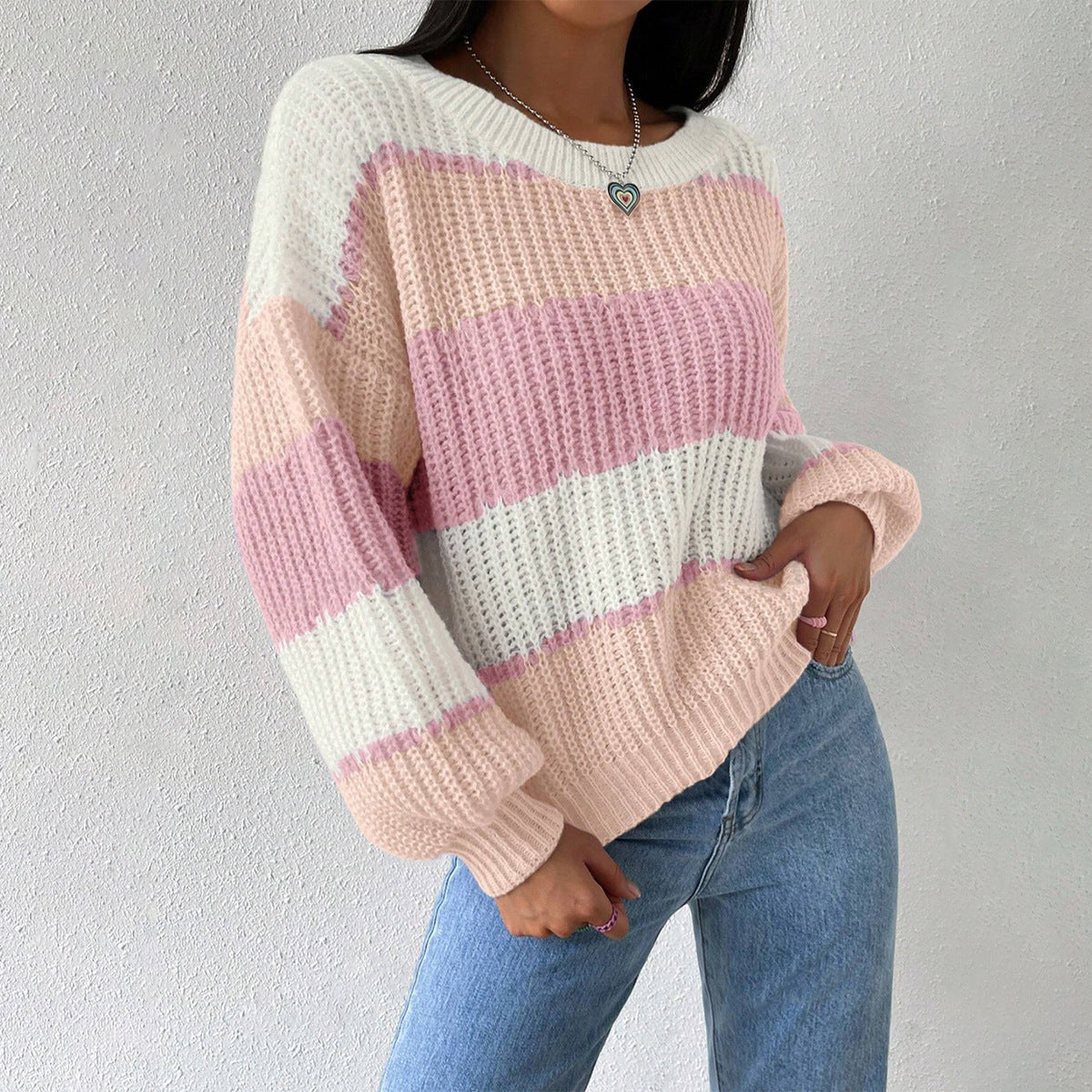Femme portant un pull rayé rose et blanc, manches longues, col rond, avec un collier cœur, posant contre un mur blanc. Mode décontractée automne.