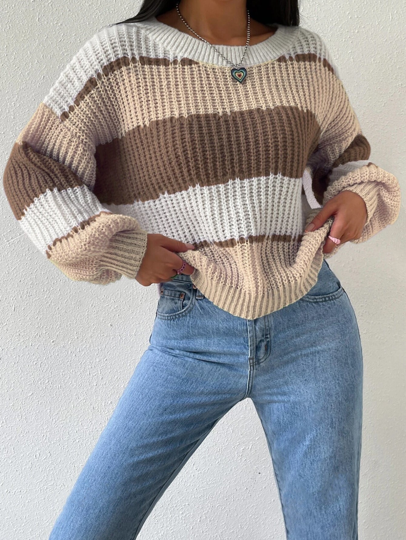 Femme portant un pull rayé beige et marron, col rond, manches longues, avec un collier pendentif, associée à un jean bleu clair ajusté.