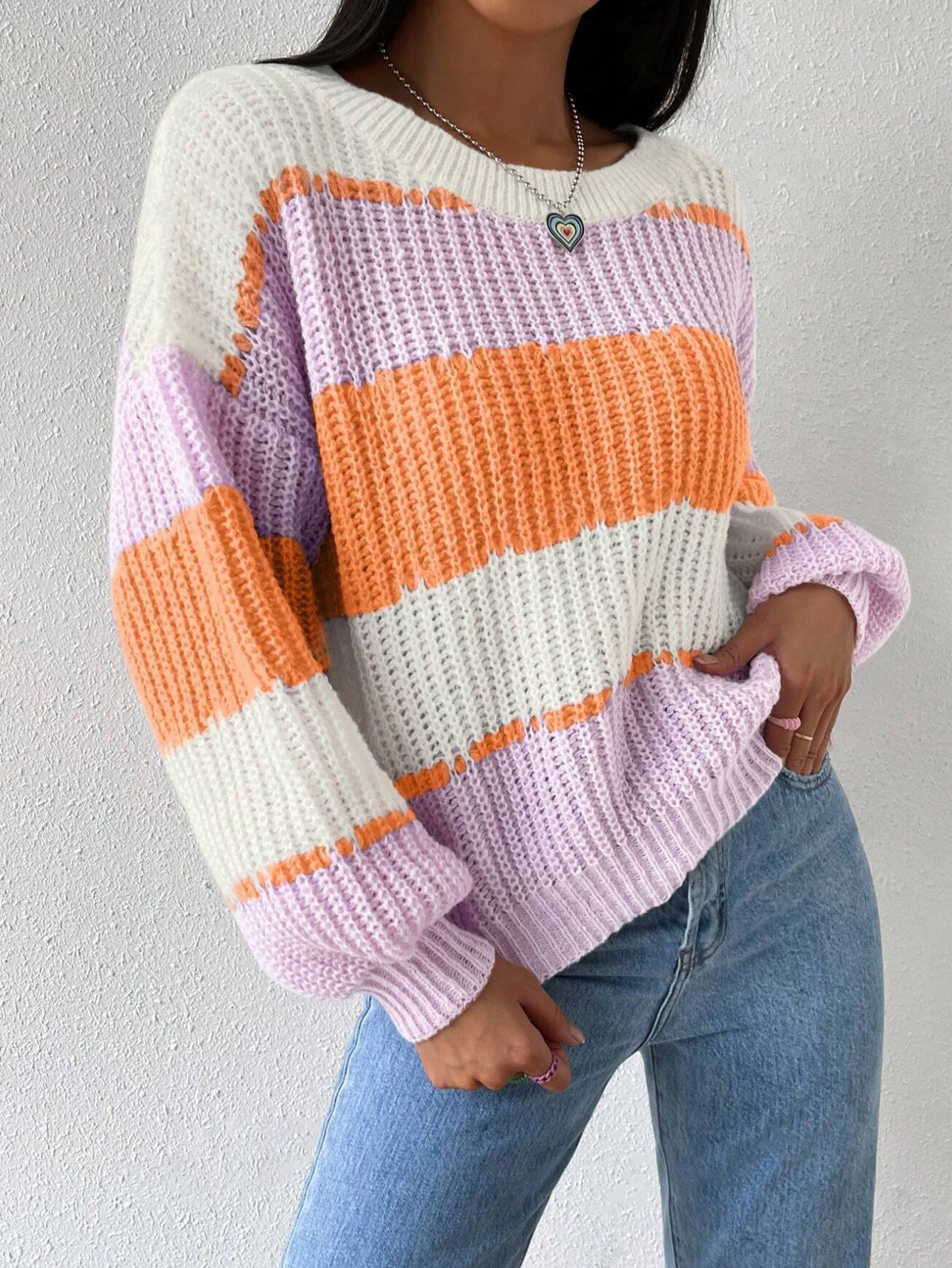Femme portant un pull tricoté à rayures orange, blanc et lilas, avec un jean bleu. Mode automne-hiver, style décontracté et tendance.