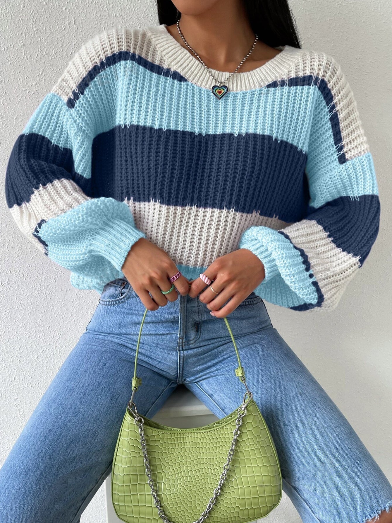 Femme portant un pull rayé bleu et blanc, tenant un sac à main vert avec chaîne argentée, assise en jean clair. Mode automne, style décontracté.
