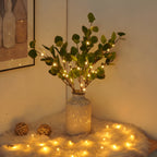 Vase en céramique avec branches de feuillage vert et guirlandes lumineuses LED, posé sur un tapis moelleux, créant une ambiance chaleureuse et cosy.