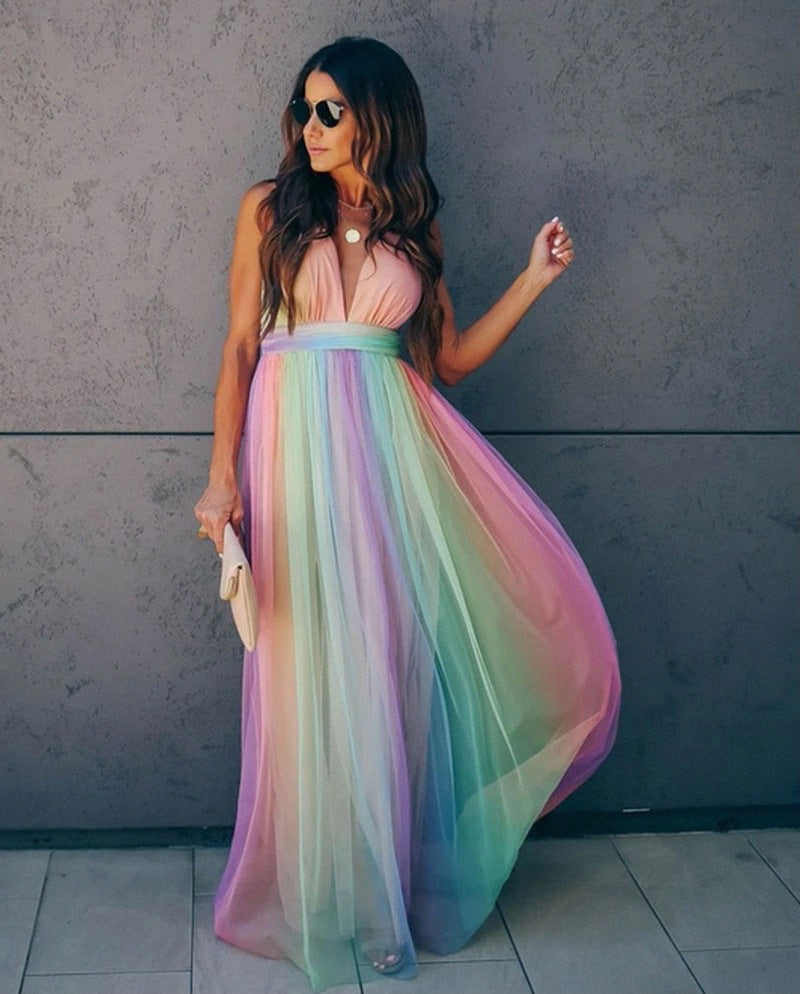 Femme en robe longue arc-en-ciel, lunettes de soleil, tenant une pochette beige, posant devant un mur gris. Mode élégante et colorée.