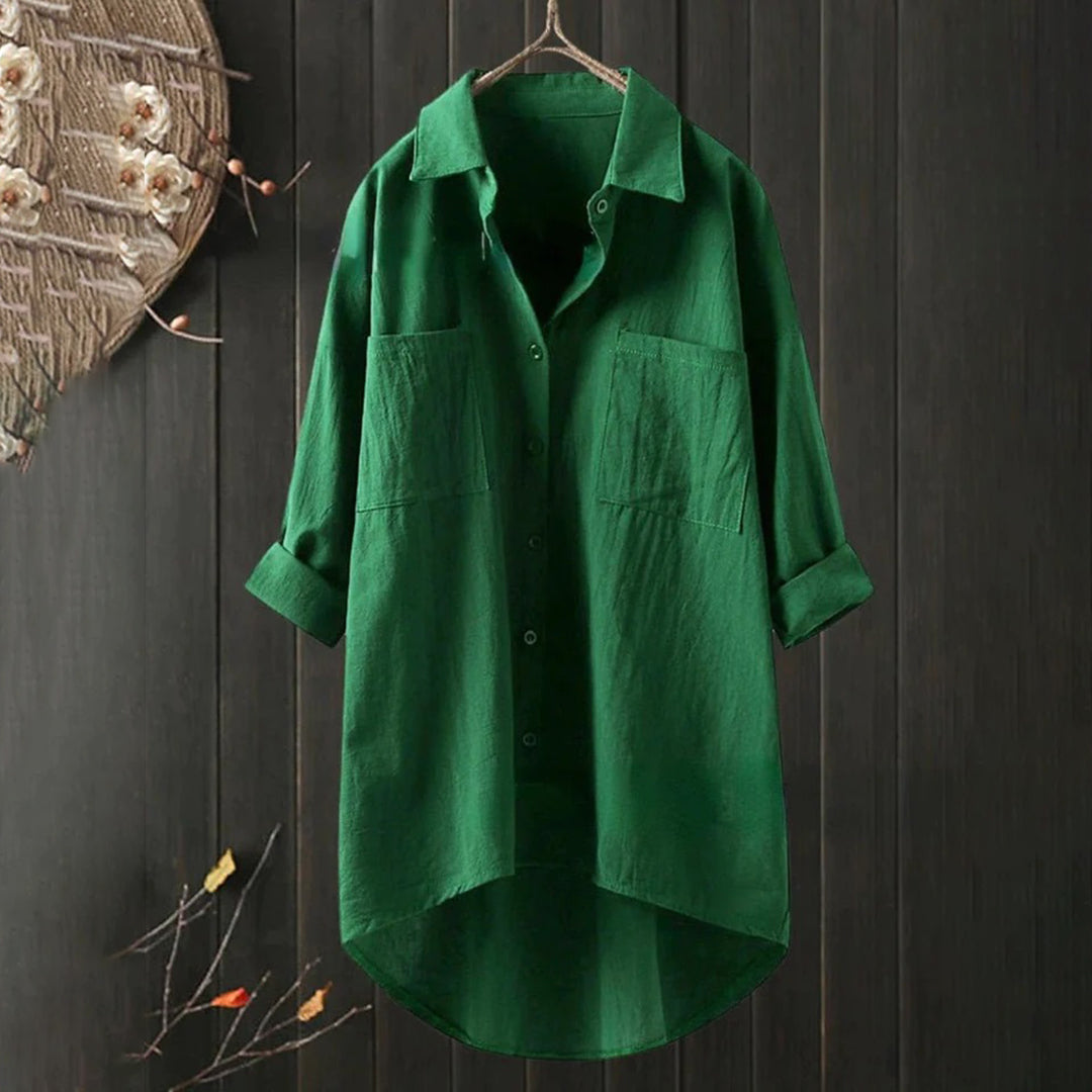 Chemise verte en lin suspendue, manches longues, col classique, boutons, poches avant, fond en bois sombre, décoration florale à gauche.