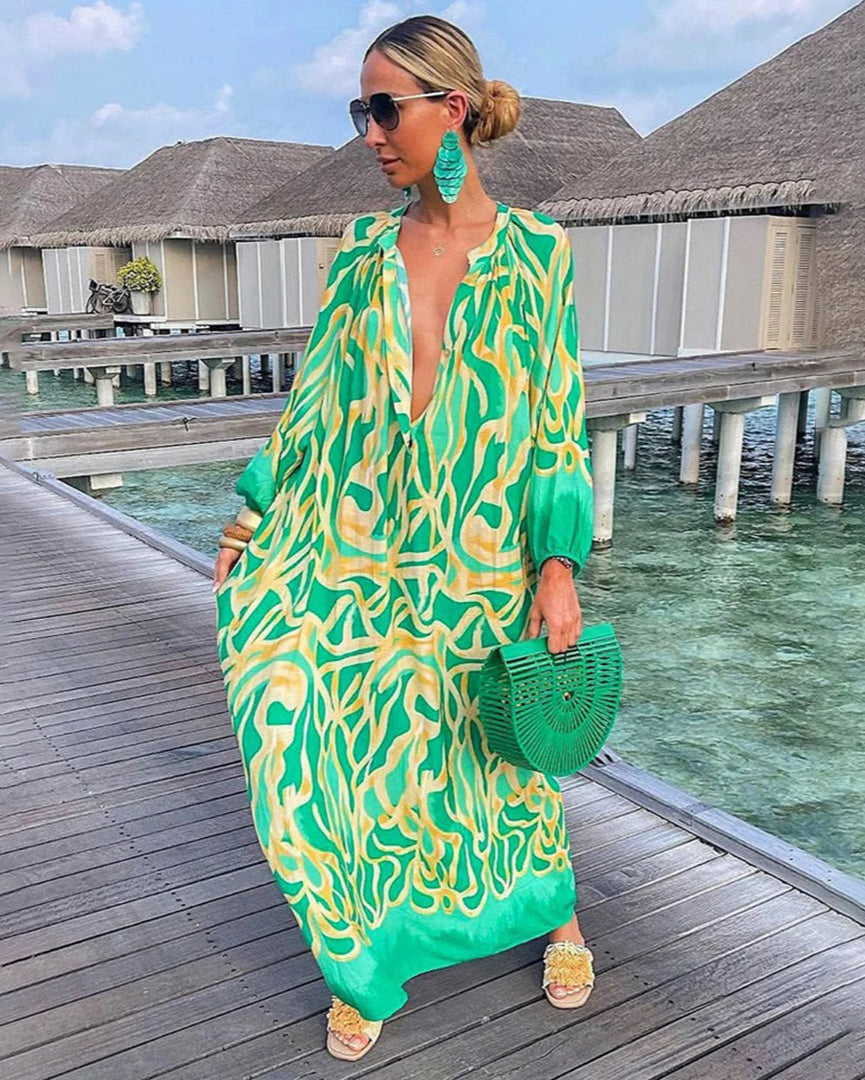 Femme en robe longue verte à motifs, portant des lunettes de soleil et des boucles d'oreilles, sur une passerelle en bois près de bungalows sur l'eau.