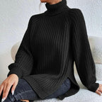 Femme portant un pull noir à col roulé en maille épaisse, manches longues, assise sur un canapé blanc. Mode automne-hiver, style décontracté.