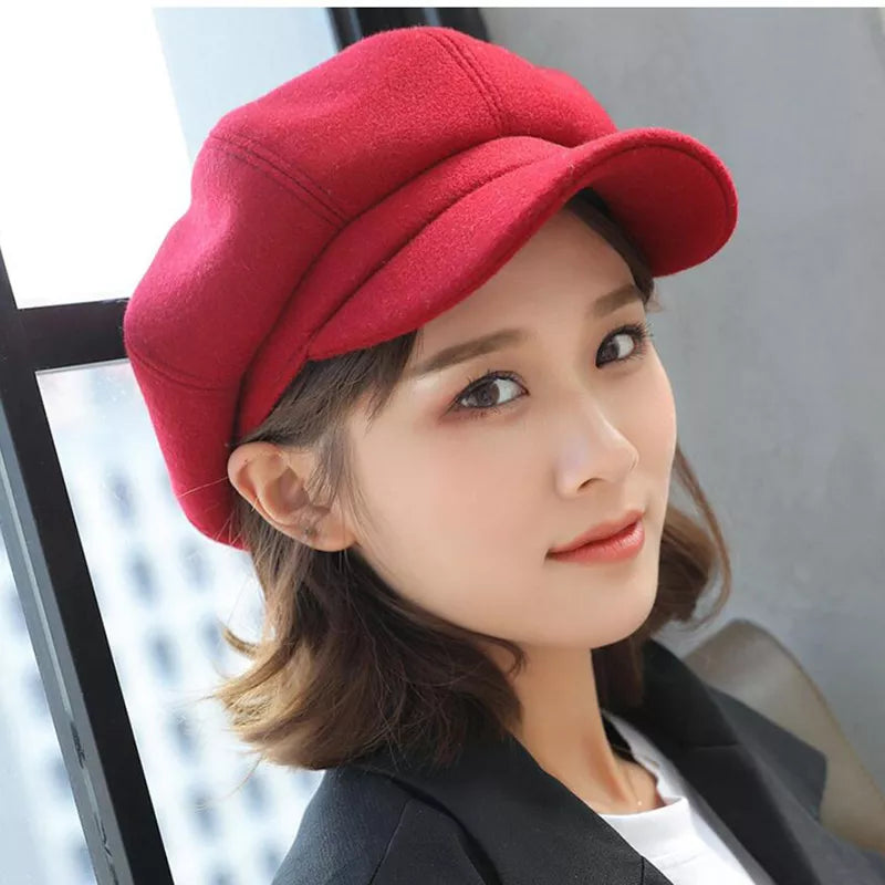 Femme portant une casquette gavroche rouge, style élégant, mode automne-hiver, accessoire tendance, devant une fenêtre.
