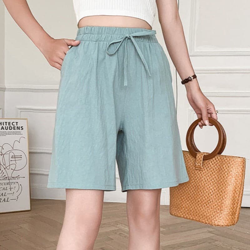 Femme portant un short bleu clair taille haute avec ceinture, tenant un sac en osier. Mode estivale, tenue décontractée, style moderne.